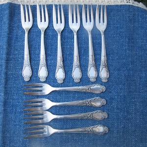 Può includere: Una collezione di forchette d'argento antiche disposte su una tovaglia blu. Le forchette hanno manici ornati con un design decorativo. L'immagine include sia forchette da tavola che forchette da dessert più piccole, mostrando un set completo per un'esperienza culinaria formale.