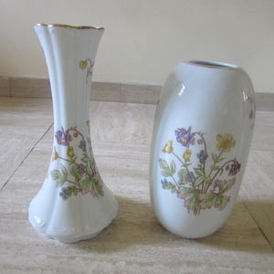 Puede incluir: Dos jarrones de porcelana blanca con delicadas ilustraciones florales en tonos amarillo, morado y verde. El jarrón más alto tiene un cuello acanalado y acampanado con borde dorado, mientras que el jarrón ovalado más corto tiene un cuerpo liso.