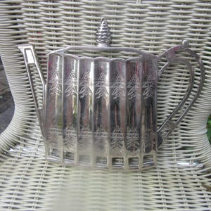 Può includere: Una teiera color argento con corpo scanalato e coperchio decorativo. La teiera presenta un manico e un beccuccio ornati ed è appoggiata su una sedia di vimini bianca. La teiera presenta disegni incisi lungo il corpo.