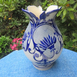 Puede incluir: Un jarrón de cerámica con una base gris claro y intrincados diseños florales azules. El jarrón tiene un cuerpo ancho y redondeado y un borde acampanado en forma de pétalo. El jarrón está sobre una superficie azul.