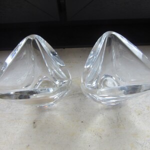 May include: Two clear glass candle holders with a triangular shape. The holders are made of thick glass and have a smooth, polished finish.