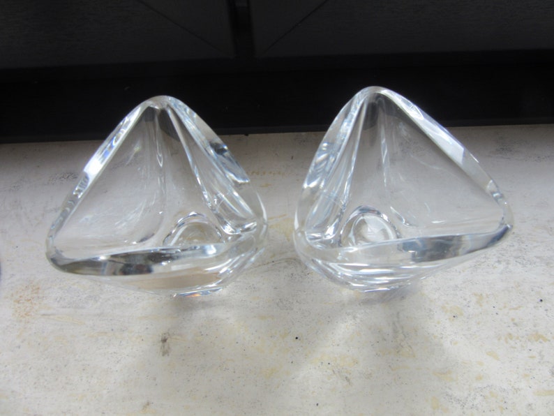 May include: Two clear glass candle holders with a triangular shape. The holders are made of thick glass and have a smooth, polished finish.