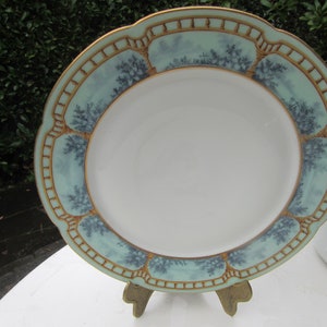 Pode incluir: Um prato de cerâmica branco com uma borda decorativa azul clara e dourada. A borda apresenta um design repetido de árvores e elementos arquitetônicos. O prato é exibido em um pequeno cavalete de madeira.