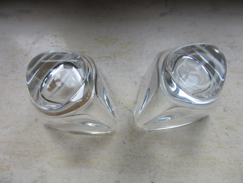 May include: Two clear glass candle holders with a unique, faceted design. The holders are empty and have a reflective surface.