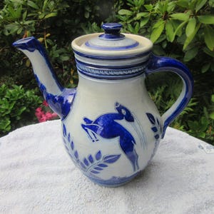 May include: A ceramic teapot with a lid, spout, and handle. The teapot is primarily white with blue accents, including a blue handle, spout, and decorative patterns. A blue deer design is featured on the body. The teapot is sitting on a white surface.