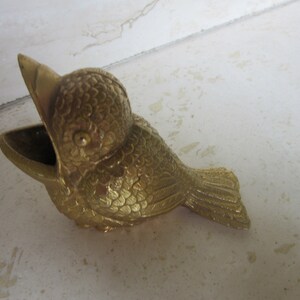Puede incluir: Un recipiente decorativo dorado con forma de p&aacute;jaro, con un pico abierto y una peque&ntilde;a abertura en forma de cuenco. El p&aacute;jaro tiene texturas de plumas detalladas y est&aacute; posicionado sobre una superficie clara. El recipiente es probablemente de metal.