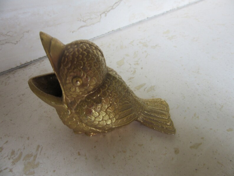 Puede incluir: Un recipiente decorativo dorado con forma de p&aacute;jaro, con un pico abierto y una peque&ntilde;a abertura en forma de cuenco. El p&aacute;jaro tiene texturas de plumas detalladas y est&aacute; posicionado sobre una superficie clara. El recipiente es probablemente de metal.
