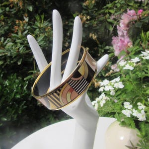 May include: A black and gold bangle bracelet with geometric patterns in shades of beige and brown is displayed on a white hand-shaped jewelry holder. The bracelet has a gold rim and is set against a backdrop of green foliage and white flowers.