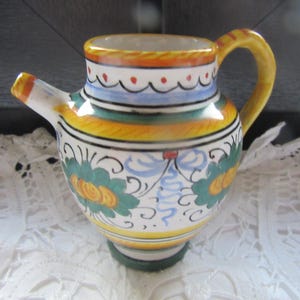 May include: A small, white ceramic pitcher with a handle and a spout. The pitcher is decorated with a colorful floral pattern in shades of yellow, green, blue, and red.