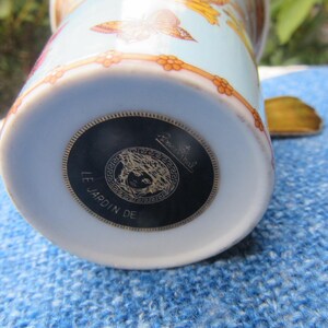 May include: A light blue and white ceramic mug with a floral and butterfly design. The bottom of the mug features a black and gold medallion with the text "LE JARDIN DE".