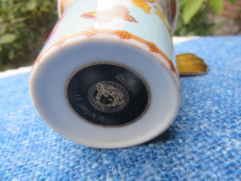 May include: A light blue and white ceramic mug with a floral and butterfly design. The bottom of the mug features a black and gold medallion with the text "LE JARDIN DE".