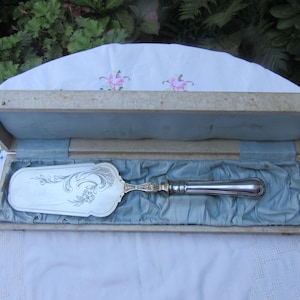Puede incluir: Una espátula para pastel plateada con diseños florales y de volutas ornamentados, presentada en una caja de madera vintage forrada con tela azul. El mango de la espátula está pulido y la caja está abierta, revelando la espátula.