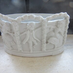 Può includere: Vaso da fiori in porcellana bianca con un design in rilievo di bambini che si tengono per mano e una ghirlanda di rose.