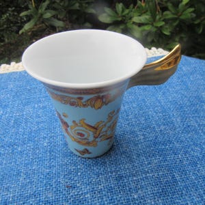 May include: A light blue ceramic teacup with a gold handle and ornate gold and brown detailing. The cup has a white interior and is set on a blue surface. The teacup is likely part of a tea set.