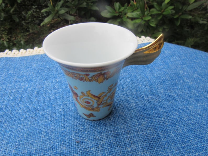 May include: A light blue ceramic teacup with a gold handle and ornate gold and brown detailing. The cup has a white interior and is set on a blue surface. The teacup is likely part of a tea set.