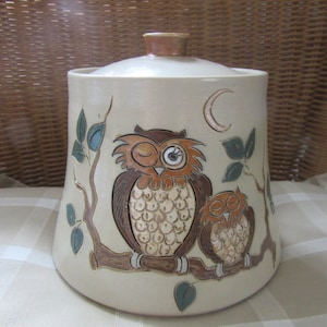 May include: Ceramic cookie jar with a cream-coloured base and a brown lid. The jar features a hand-painted design of two owls perched on a branch with green leaves. The larger owl has one eye closed and the smaller owl has both eyes closed. The jar has a crescent moon design on the side.