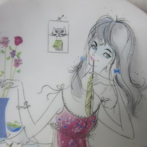 Op de afbeelding: Een witte keramische plaat met een gestileerde illustratie van een vrouw in een roze jurk, die een veer vasthoudt. De plaat heeft een vaas met bloemen en een kattenkalender. Het kunstwerk is uitgevoerd in een minimalistische stijl met zwarte contouren en pastelkleuren.