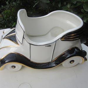 Może przedstawiać: Biała ceramiczna doniczka w kształcie zabytkowego samochodu z czarnymi i złotymi akcentami. Doniczka ma zaokrąglony kształt i szeroki otwór.