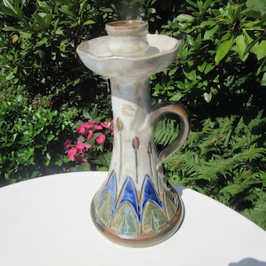 Puede incluir: Un candelero de cerámica con asa. El candelero es de color blanquecino con detalles azules, verdes y marrones. La base tiene un diseño acampanado con formas azules y verdes. El asa es marrón.