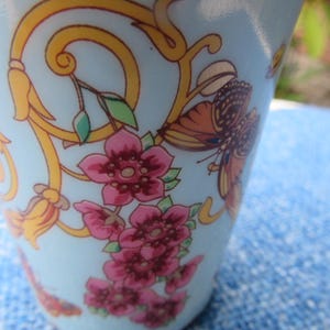 May include: A light blue ceramic cup with a floral and butterfly design. The cup features pink flowers, orange and brown butterflies, and gold scrollwork. The design is set against a light blue background.
