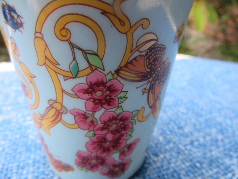 May include: A light blue ceramic cup with a floral and butterfly design. The cup features pink flowers, orange and brown butterflies, and gold scrollwork. The design is set against a light blue background.