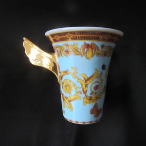 May include: A decorative ceramic mug with a light blue background and gold baroque-style patterns. The rim has a brown and gold border. The handle is gold and shaped like a wing. The mug is likely for coffee or tea.