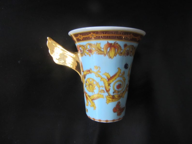 May include: A decorative ceramic mug with a light blue background and gold baroque-style patterns. The rim has a brown and gold border. The handle is gold and shaped like a wing. The mug is likely for coffee or tea.