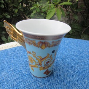May include: A light blue ceramic mug with a gold-colored butterfly wing handle. The mug features a decorative pattern in gold and brown, with a white interior. The mug is sitting on a blue surface.