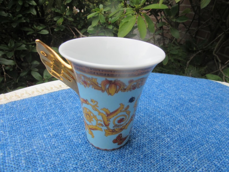 May include: A light blue ceramic mug with a gold-colored butterfly wing handle. The mug features a decorative pattern in gold and brown, with a white interior. The mug is sitting on a blue surface.