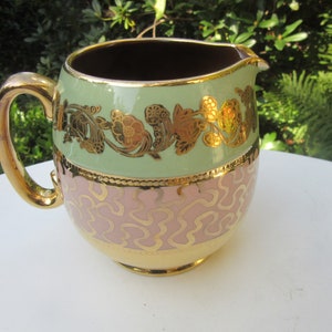 Può includere: Un'antica brocca in ceramica con un design floreale verde e rosa e dettagli dorati. La brocca ha un corpo rotondo e un manico.
