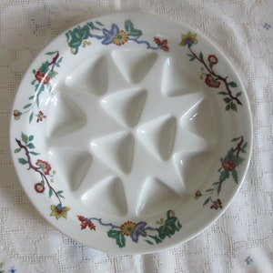 Puede incluir: Un plato de cerámica blanco para caracoles con doce compartimentos triangulares. El borde está decorado con un estampado floral en rojo, amarillo, verde y azul. El plato está diseñado para servir caracoles.