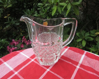 Puede incluir: Una jarra de cristal transparente con asa y boquilla. La jarra tiene un patrón de diamantes en la mitad inferior y está sobre un mantel a cuadros rojo y blanco. El fondo presenta follaje verde y flores rosas.