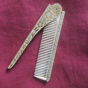 May include: A folding pocket comb featuring a metallic handle with an intricate gold-toned floral and geometric engraving. The comb teeth are white and made of a smooth material. The handle is hinged to allow the comb to fold into the casing.