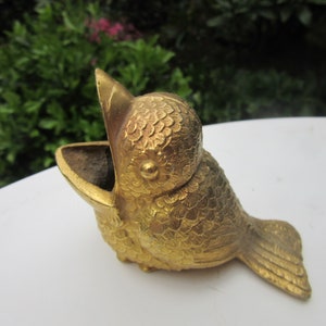 Puede incluir: Un recipiente dorado con forma de p&aacute;jaro y pico abierto. El p&aacute;jaro tiene texturas detalladas en forma de escamas que cubren su cuerpo y una peque&ntilde;a cola. El recipiente se utiliza probablemente para guardar objetos peque&ntilde;os o como pieza decorativa.
