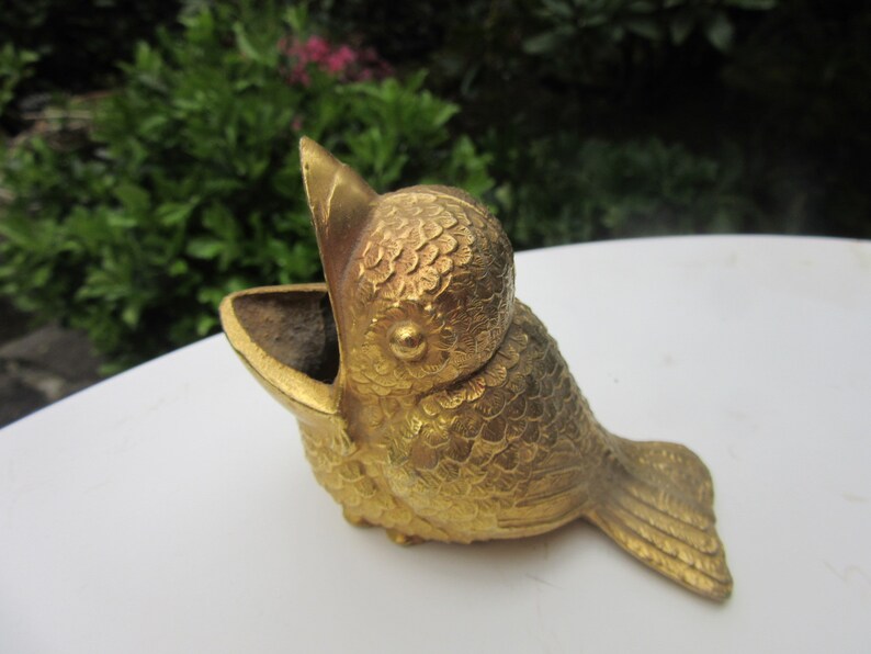 Puede incluir: Un recipiente dorado con forma de p&aacute;jaro y pico abierto. El p&aacute;jaro tiene texturas detalladas en forma de escamas que cubren su cuerpo y una peque&ntilde;a cola. El recipiente se utiliza probablemente para guardar objetos peque&ntilde;os o como pieza decorativa.