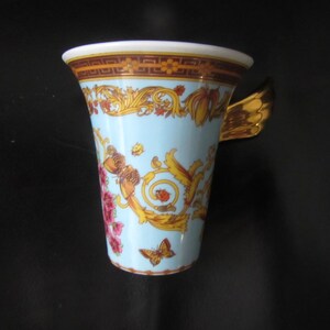 May include: A light blue ceramic mug with a gold butterfly wing handle. The mug features a decorative design with gold, red, and orange floral and scrollwork patterns. A butterfly is depicted on the front of the mug.