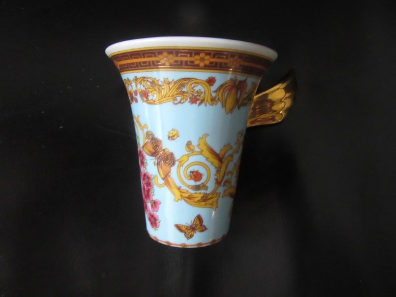 May include: A light blue ceramic mug with a gold butterfly wing handle. The mug features a decorative design with gold, red, and orange floral and scrollwork patterns. A butterfly is depicted on the front of the mug.