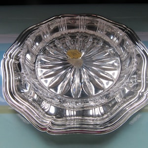 Op de afbeelding: Een verzilverde serveerschaal met een heldere, facetgeslepen glazen inzet. De schaal heeft een geschulpte rand en een stervormig patroon in het midden. Een kleine, goudkleurige sticker is zichtbaar in het midden van het glas.