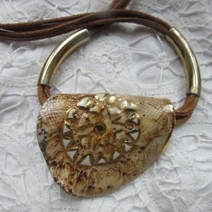 May include: A brown leather cord necklace with a gold metal ring and a large, brown, shell pendant with a white and gold design.