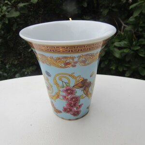 May include: A decorative ceramic vase with a light blue background, adorned with gold and brown ornate patterns, and pink floral accents. The vase has a white rim and a flared shape, suitable for displaying flowers.