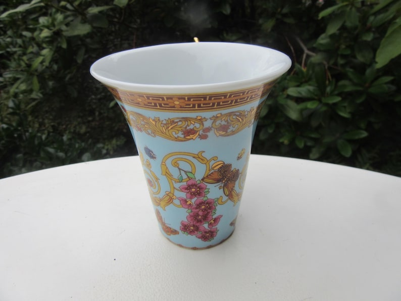 May include: A decorative ceramic vase with a light blue background, adorned with gold and brown ornate patterns, and pink floral accents. The vase has a white rim and a flared shape, suitable for displaying flowers.