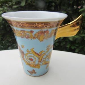 May include: A light blue ceramic mug with a gold handle and ornate gold and orange floral designs. The rim has a gold and brown geometric pattern. A butterfly is visible on the side of the mug.