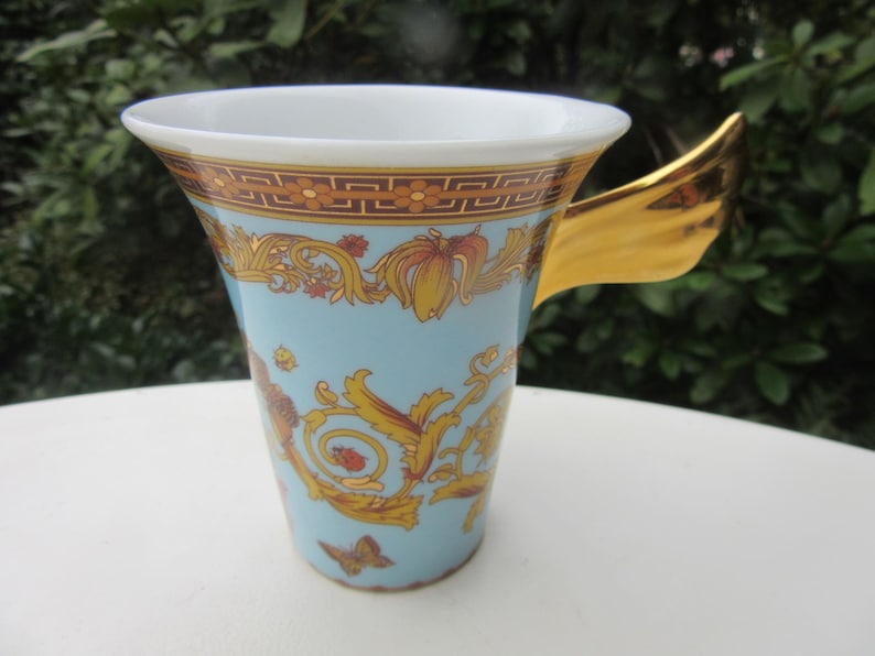 May include: A light blue ceramic mug with a gold handle and ornate gold and orange floral designs. The rim has a gold and brown geometric pattern. A butterfly is visible on the side of the mug.