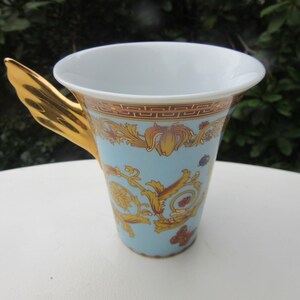 May include: A decorative ceramic cup with a light blue background and gold accents. The cup features a gold handle and ornate gold floral designs. The rim of the cup has a gold and brown border.