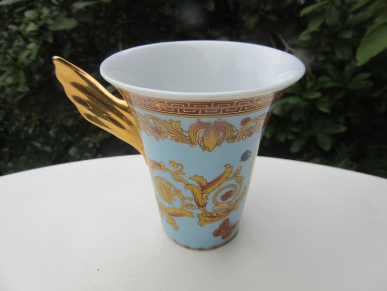 May include: A decorative ceramic cup with a light blue background and gold accents. The cup features a gold handle and ornate gold floral designs. The rim of the cup has a gold and brown border.