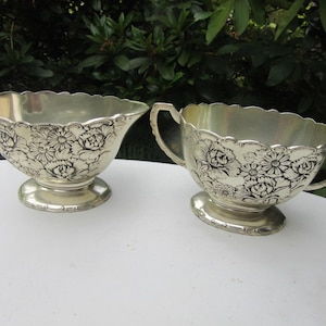 Puede incluir: Dos fuentes plateadas con diseños florales. Una tiene un pico y un asa, la otra tiene dos asas. Ambas tienen bordes festoneados y se asientan sobre una base redonda. El patrón floral es en blanco y negro.