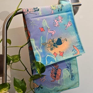 Puede incluir: Dos toallas de mano estampadas y coloridas colgadas de una barra plateada. Las toallas presentan un fondo azul, morado y turquesa con ilustraciones de dibujos animados de criaturas marinas y personajes. Una toalla tiene la palabra "Tropikaldaize" impresa.