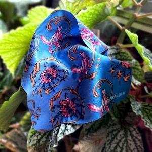 Może przedstawiać: Złożony niebieski jedwabny szalik z powtarzającym się wzorem różowych i niebieskich owadów, brązowych liści i gwiazd. Szalik ma nadrukowany tekst "WIKALDALE". Szalik jest otoczony zielonymi liśćmi.