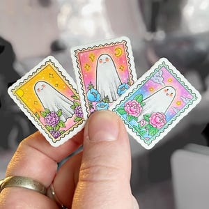 以下が含まれることがあります： 遊び心のあるデザインのカラフルなゴーストステッカー3枚。各ステッカーには、笑顔の白いゴーストが描かれており、花と星で囲まれています。ステッカーは、波状の縁取りのある白い背景に印刷されています。