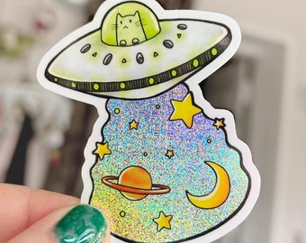 Könnte beinhalten: Ein holografischer Aufkleber mit einer Cartoon-Katze, die in einer fliegenden Untertasse sitzt. Die Untertasse fliegt über einem glitzernden blauen Hintergrund mit Sternen und einer Mondsichel.